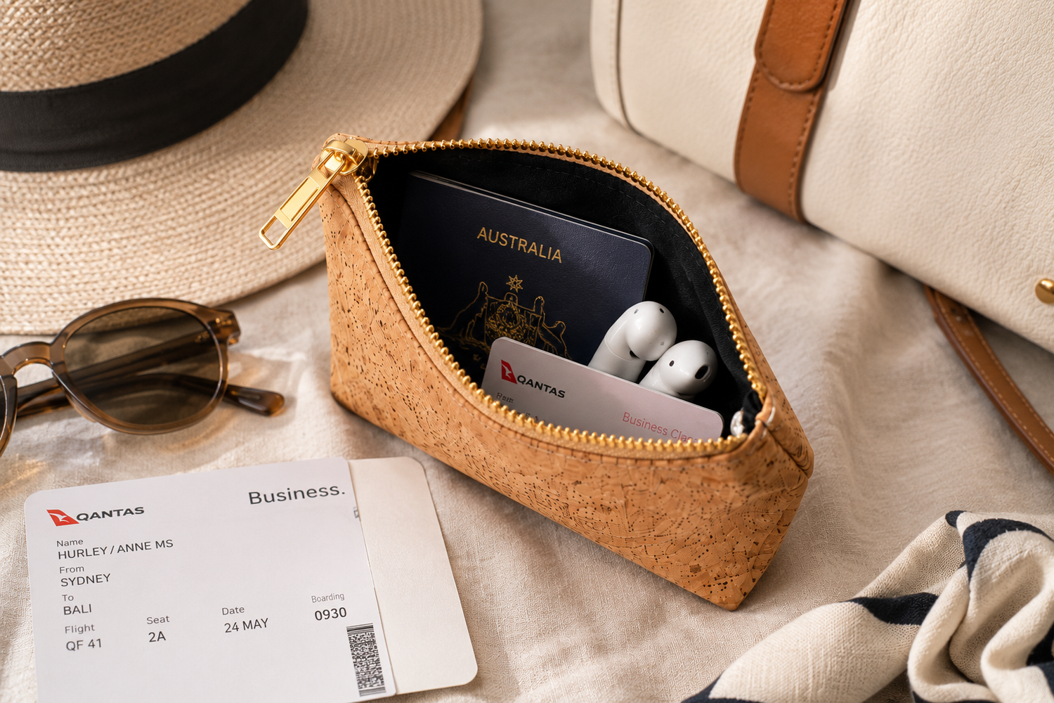 A small zippered pouch made of natural cork with a gold zipper, shown open and filled with travel items including an Australia passport, white earbuds, and a Qantas card. It is styled on a light fabric surface with a straw hat, sunglasses, a boarding pass, and part of a cream handbag nearby.